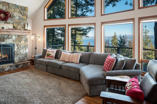 Cozy living room with a large gray sectional, fireplace, and stunning lake view through tall windows. Christmas decor visible.